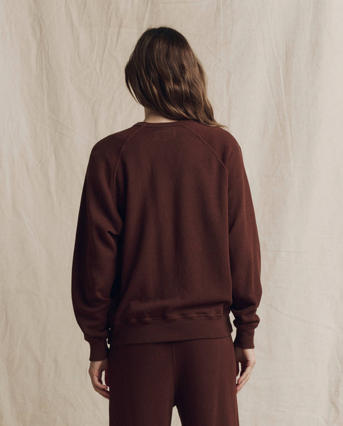 Wearing THE GREAT. The College Sweatshirt. Solid -- Driftwood from the F26 SEASONAL CORE collection, a person with long brown hair stands with their back to the camera against a beige fabric backdrop, embodying a timeless look handcrafted in Los Angeles.