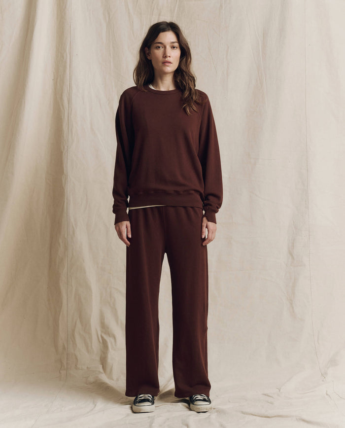 A woman stands against a beige fabric backdrop wearing THE GREAT.’s The College Sweatshirt in Driftwood, paired with matching pants and black and white sneakers.