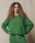 A person with curly hair wears THE GREAT.’s The College Sweatshirt. Solid -- Fennel from the SU26 SEASONAL CORE collection, paired with matching pants, standing hands on hips against a neutral fabric backdrop.