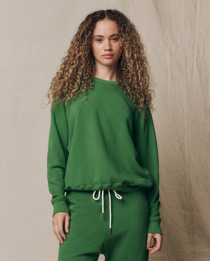 A person with curly hair wears THE GREAT. SU26 SEASONAL CORE The College Sweatshirt, Solid -- Fennel, and matching sweatpants against a beige fabric backdrop.