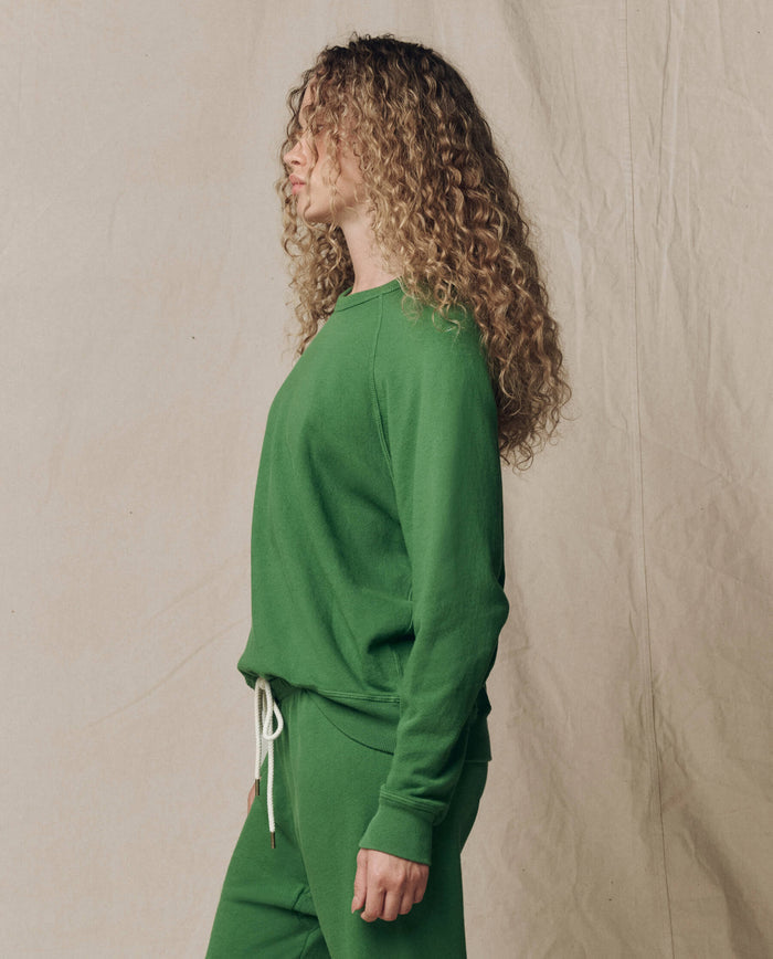 A person with curly hair stands in profile wearing THE GREAT.'s The College Sweatshirt in Solid Fennel (SU26 SEASONAL CORE) and matching pants, set against a plain, light fabric backdrop.