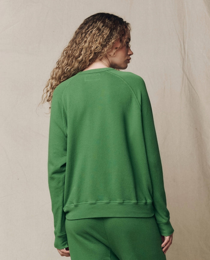 Person with curly hair wears THE GREAT.'s The College Sweatshirt in Solid Fennel, SU26 SEASONAL CORE, and matching pants, standing with their back to the camera against a neutral backdrop.