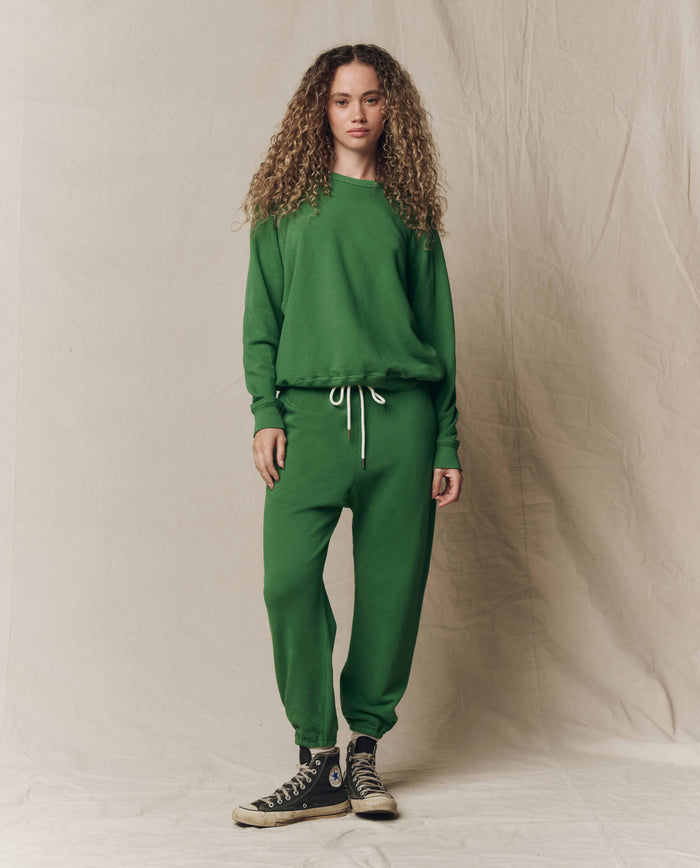 Someone with long curly hair wears THE GREAT.'s The College Sweatshirt. Solid -- Fennel with matching sweatpants, posed against a plain light backdrop.