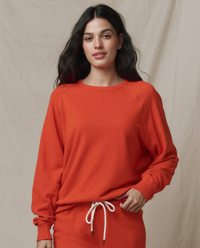 A person with long dark hair wears THE GREAT.'s The College Sweatshirt in Solid Persimmon from the SP26 SEASONAL CORE, paired with coordinating pants with a white drawstring, standing before a beige fabric backdrop.