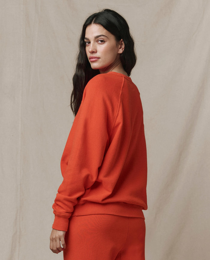 A woman with long dark hair models THE GREAT.'s The College Sweatshirt in Persimmon, standing sideways and looking at the camera against a neutral background.