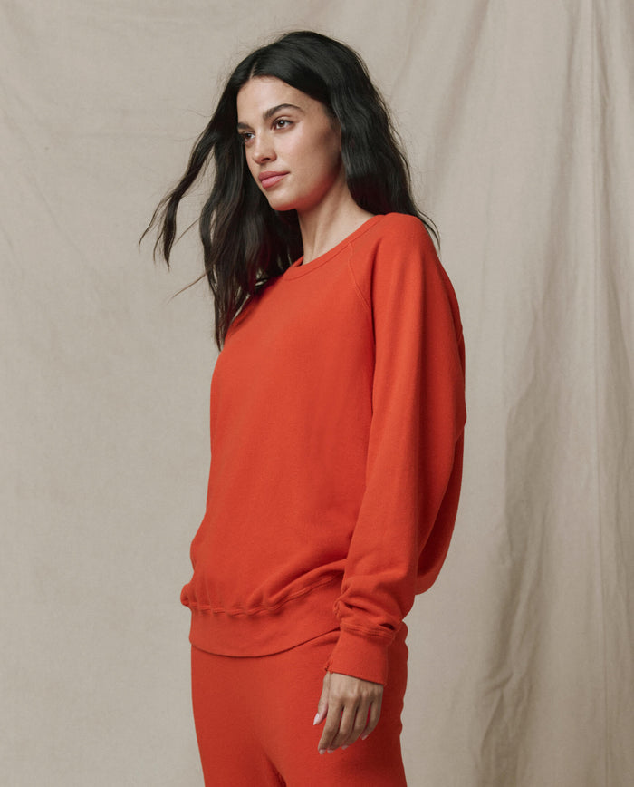 A woman stands against a beige backdrop, wearing THE GREAT.'s The College Sweatshirt in Persimmon from the SP26 SEASONAL CORE—an ode to handcrafted Los Angeles style.