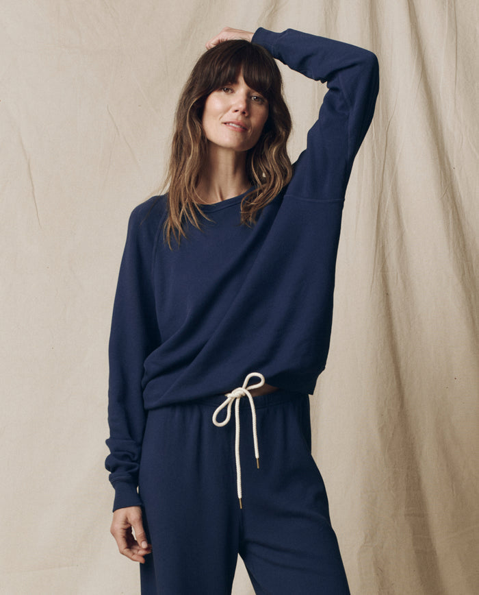 A woman models THE GREAT. FALL 23 KNITS The College Sweatshirt in True Navy, paired with matching drawstring pants, standing before a beige fabric backdrop, her hand resting on her head.