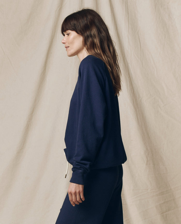 A woman with long brown hair stands in profile against a beige fabric background, wearing THE GREAT. FALL 23 KNITS The College Sweatshirt in Solid -- True Navy with matching pants.