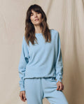 Someone wears THE GREAT. SU25 SEASONAL KNITS The College Sweatshirt, Solid -- Vista Blue, with matching pants, standing before a beige fabric backdrop.