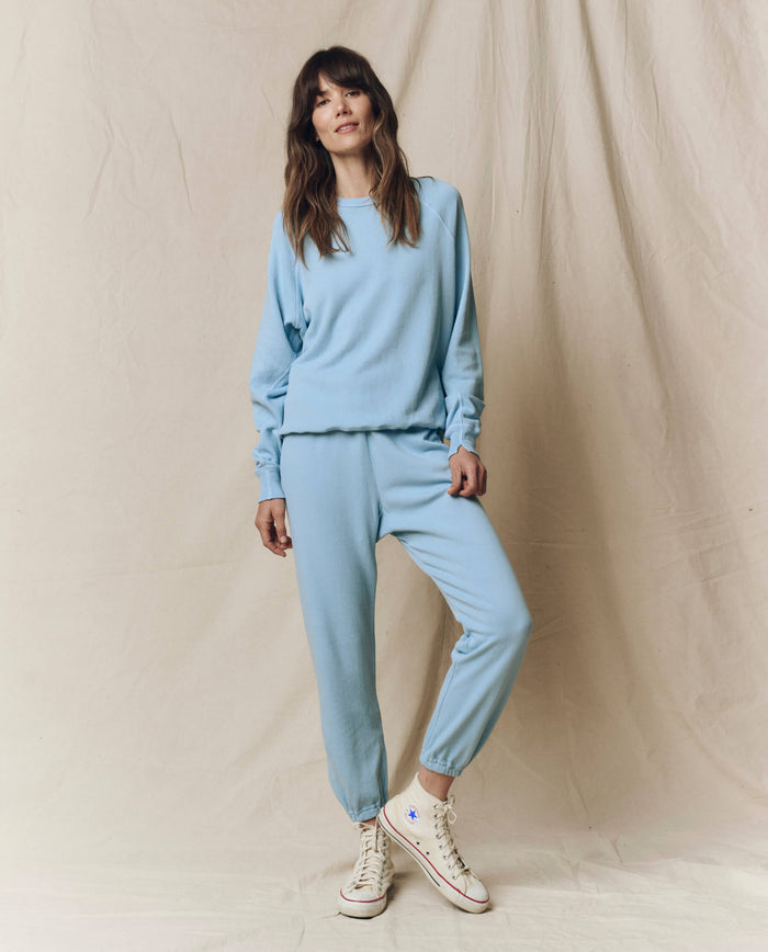 A woman stands before a beige backdrop wearing THE GREAT.'s The College Sweatshirt in Solid Vista Blue from the SU25 Seasonal Knits, matched with jogger pants and white high-top sneakers.