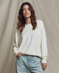 A woman with wavy brown hair wears THE GREAT. CORE KNITS The College Sweatshirt in Washed White with light blue jeans, standing before a beige fabric backdrop.