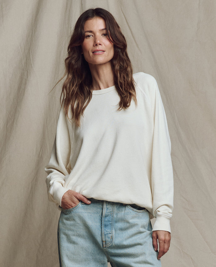 A woman with wavy brown hair wears THE GREAT. CORE KNITS The College Sweatshirt in Washed White with light blue jeans, standing before a beige fabric backdrop.