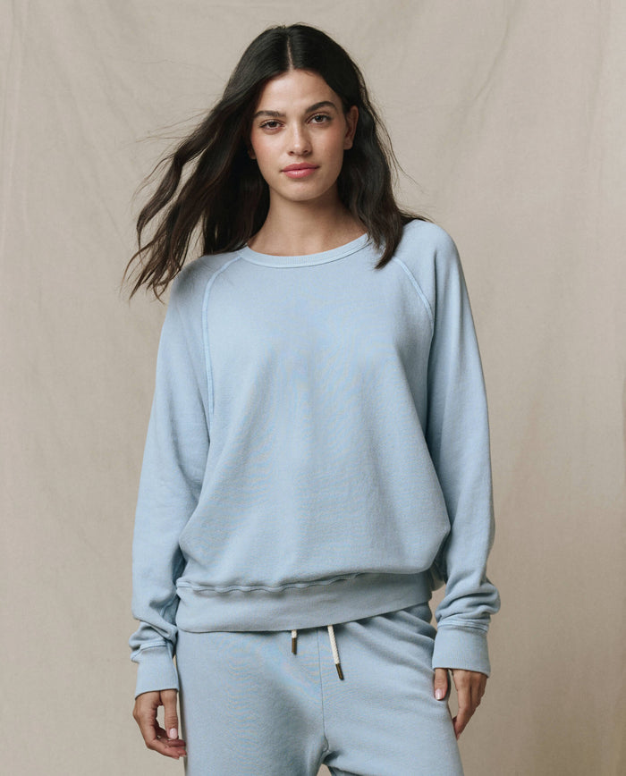 A woman models The College Sweatshirt in Whisper Blue by THE GREAT. SP26 SEASONAL CORE, paired with matching pants, standing against a plain beige background and looking at the camera with a neutral expression.