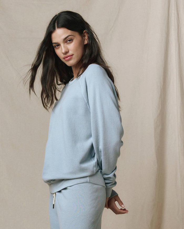 Woman with long dark hair wears THE GREAT. SP26 SEASONAL CORE The College Sweatshirt in Whisper Blue, styled for a handcrafted Los Angeles vibe, blending comfort and effortless cool against a neutral beige background.