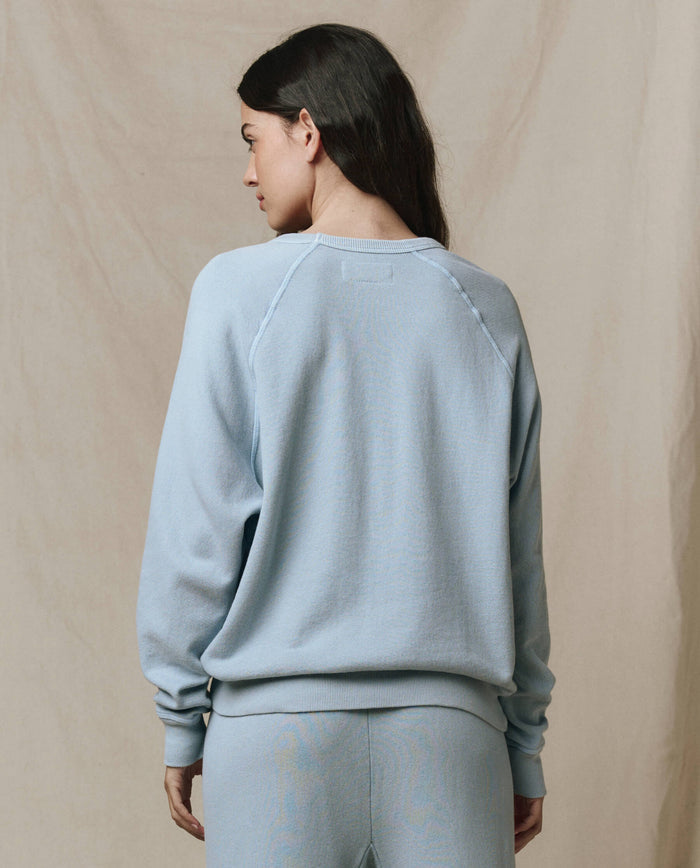 A person with long dark hair, seen from behind, wears THE GREAT. SP26 SEASONAL CORE’s The College Sweatshirt in Whisper Blue against a beige fabric backdrop.