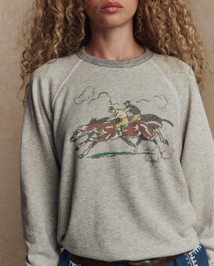A woman with curly hair wears THE GREAT. PF26’s The College Sweatshirt w/ Derby Graphic in soft heather grey, featuring a vintage illustration of two jockeys racing horses.