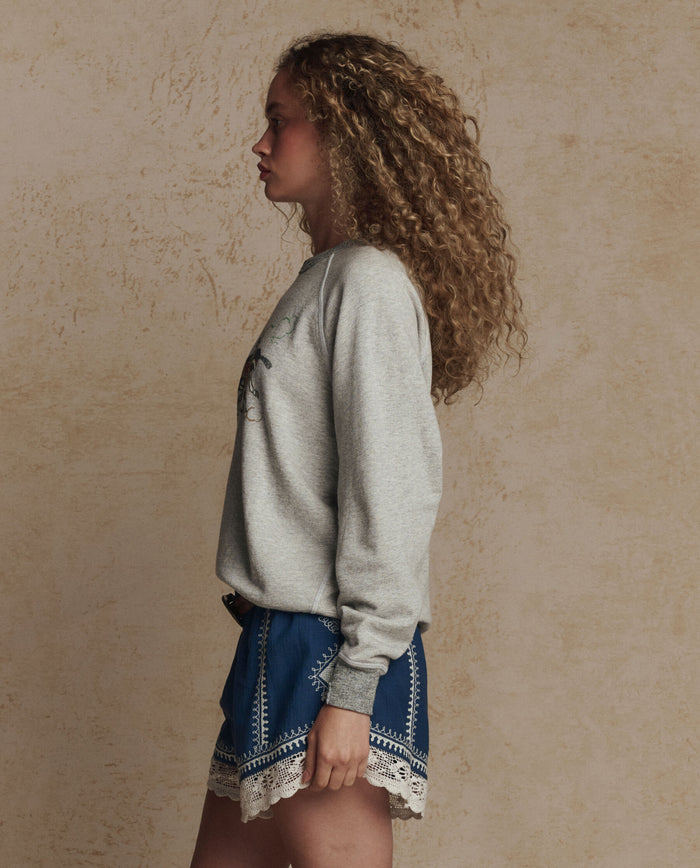 A person with curly hair stands in profile against a beige textured wall, wearing THE GREAT. PF26 The College Sweatshirt w/ Derby Graphic in soft heather grey and blue shorts with white lace trim.