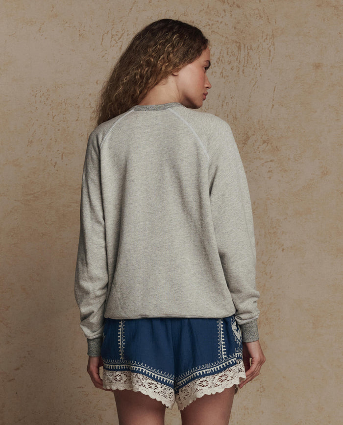 A woman with long wavy hair stands facing away, wearing THE GREAT. PF26 The College Sweatshirt w/ Derby Graphic in Soft Heather Grey and blue shorts with white lace trim, set against a textured beige background.