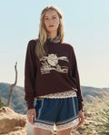 A woman stands outdoors in a desert landscape with cacti, wearing THE GREAT. PF26 The College Sweatshirt w/ Everglade Graphic in Toasted Walnut, blue shorts with white lace trim, and a leopard print collar.
