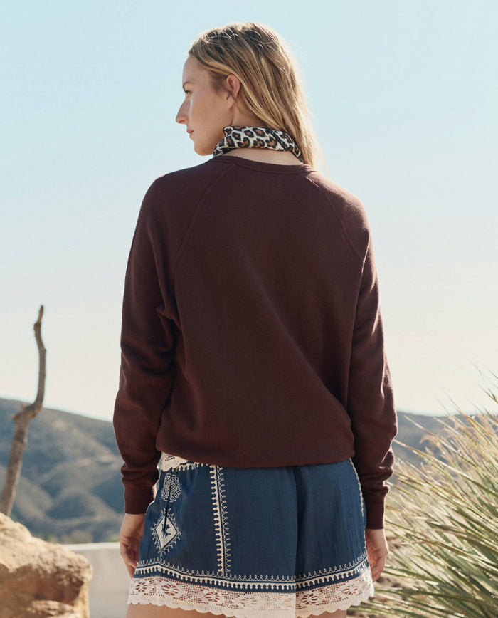 Outdoors with mountains behind, a person faces away wearing THE GREAT. PF26's The College Sweatshirt w/ Everglade Graphic in Toasted Walnut, a blue patterned skirt with white lace trim, and a leopard print scarf.