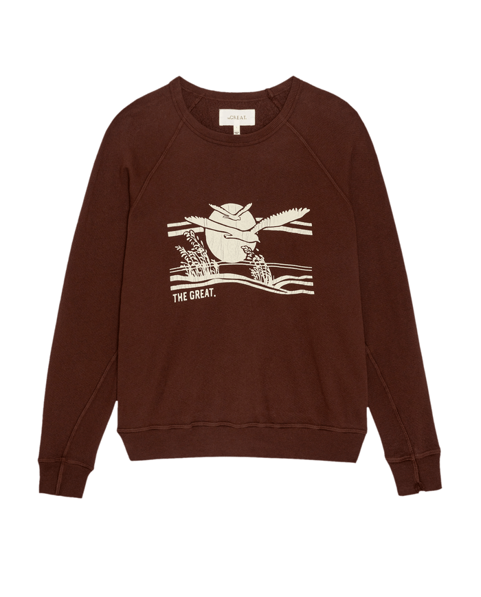 The College Sweatshirt w/ Everglade Graphic by THE GREAT. PF26 is an oversized vintage style in toasted walnut, made from double washed terry with a white flying bird over a landscape and "THE GREAT." on the front.
