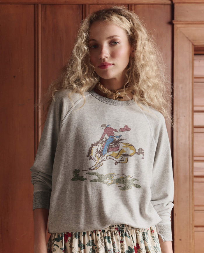 A woman with long curly blonde hair wears THE GREAT. SP26 D1 The College Sweatshirt w/ Rider Graphic in Soft Heather Grey and a floral skirt, standing before wooden paneling.