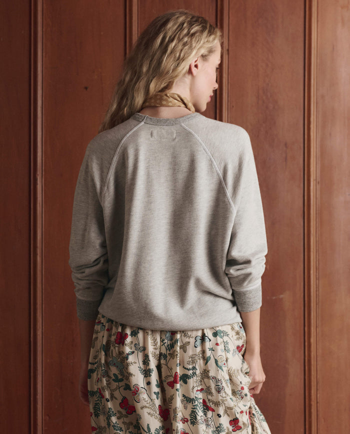 A woman with long blonde hair stands facing a wooden wall, wearing THE GREAT. SP26 D1's The College Sweatshirt w/ Rider Graphic in soft heather grey and a floral-patterned skirt.