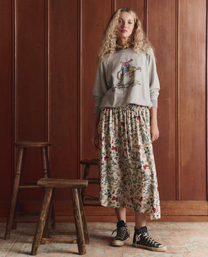 A woman stands indoors in THE GREAT. SP26 D1 The College Sweatshirt w/ Rider Graphic, Novelty—Soft Heather Grey, paired with a floral midi skirt and black sneakers; two wooden stools sit beside her against a wood-paneled wall.