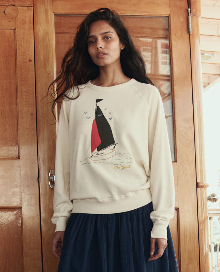 A woman stands indoors in THE GREAT. SU26 The College Sweatshirt W/ Sailing Boat Applique - Washed White and a navy skirt, set against a wooden door.