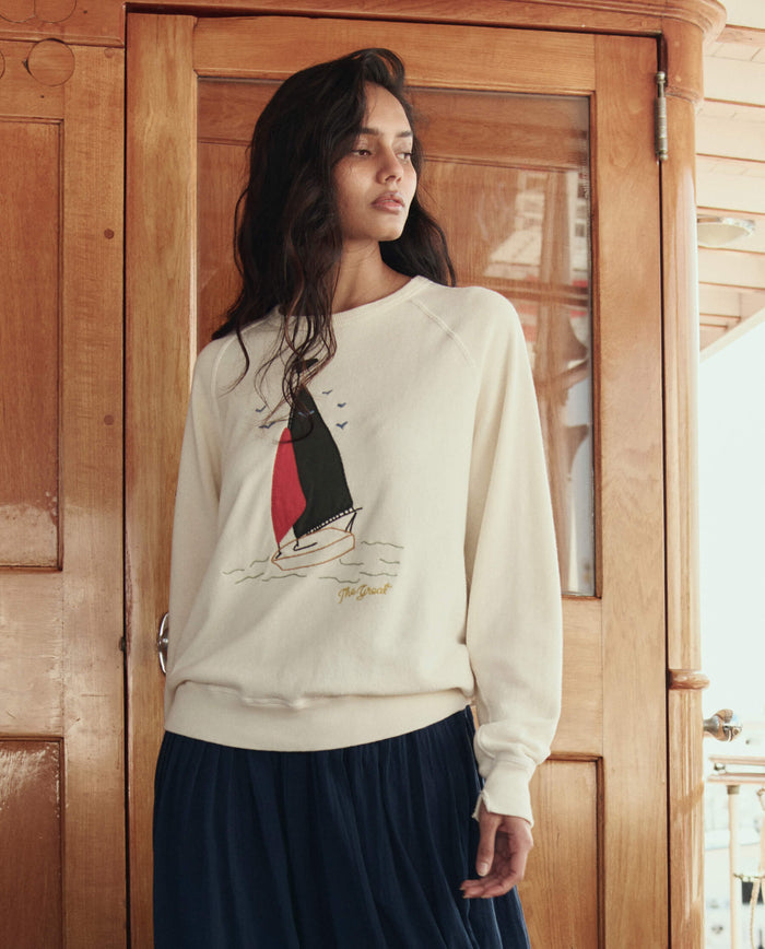 A woman stands indoors before wooden doors, wearing THE GREAT. SU26's The College Sweatshirt W/ Sailing Boat Applique in Washed White, paired with a dark blue skirt.