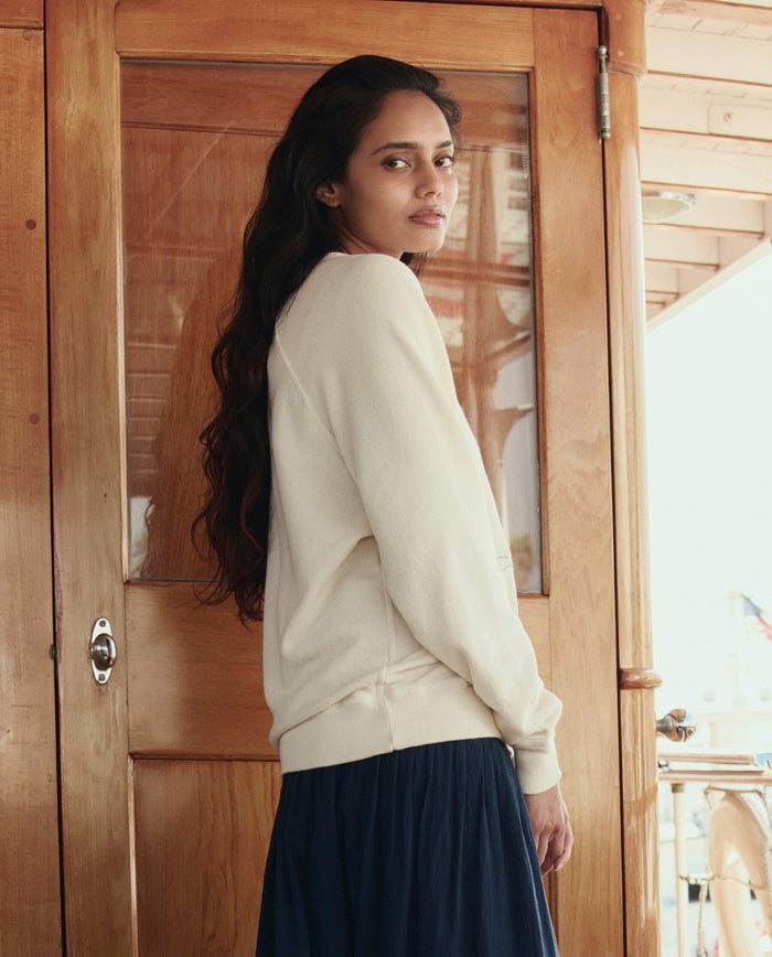 A woman with long wavy hair stands indoors near a wooden door, wearing THE GREAT. SU26 The College Sweatshirt W/ Sailing Boat Applique in Washed White and a dark skirt, looking over her shoulder at the camera.