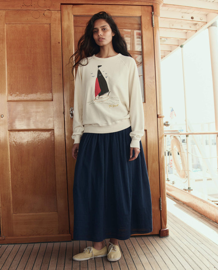 A woman stands on a wooden deck in front of a door, wearing THE GREAT. SU26 The College Sweatshirt W/ Sailing Boat Applique in washed white, paired with a navy maxi skirt and beige shoes.