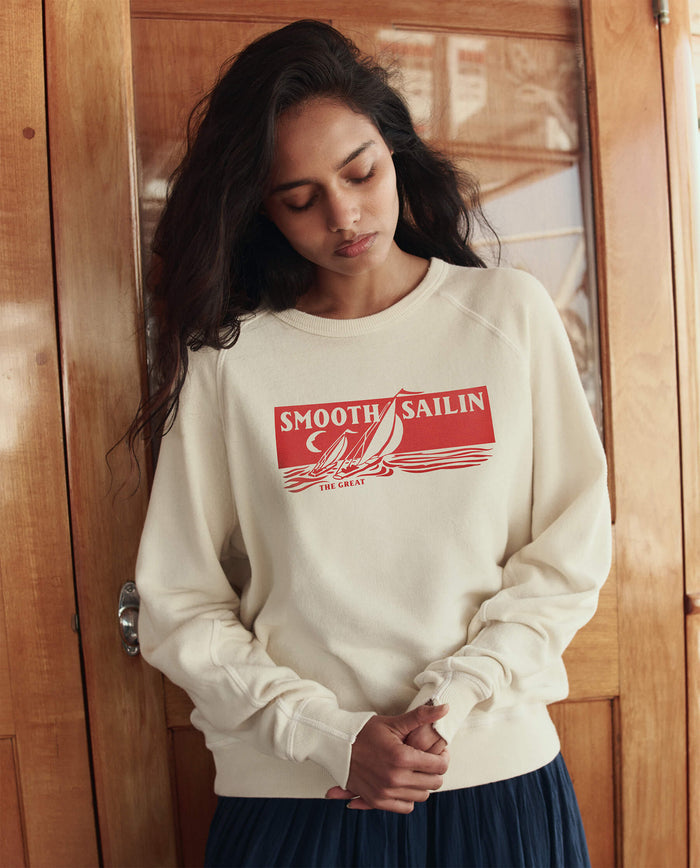 Woman with long dark hair wears THE GREAT. SU26 The College Sweatshirt W/ Smooth Sailin Graphic in Washed White, standing indoors against wooden paneling and looking down with hands clasped.