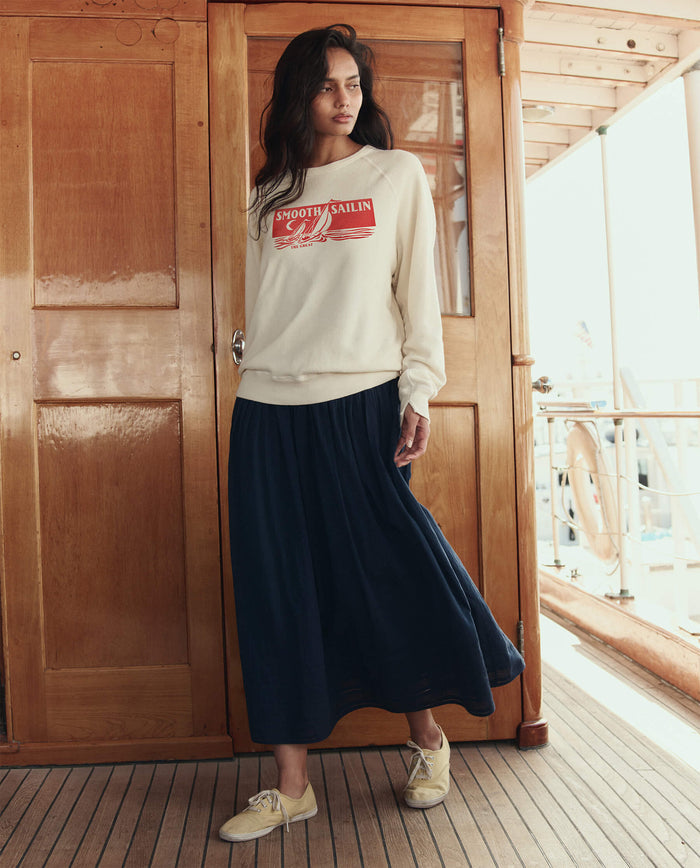 A woman stands on a wooden deck wearing THE GREAT. SU26 The College Sweatshirt w/ Smooth Sailin Graphic in washed white and a long navy skirt, paired with light shoes. Wooden doors and ship railings are in the background.