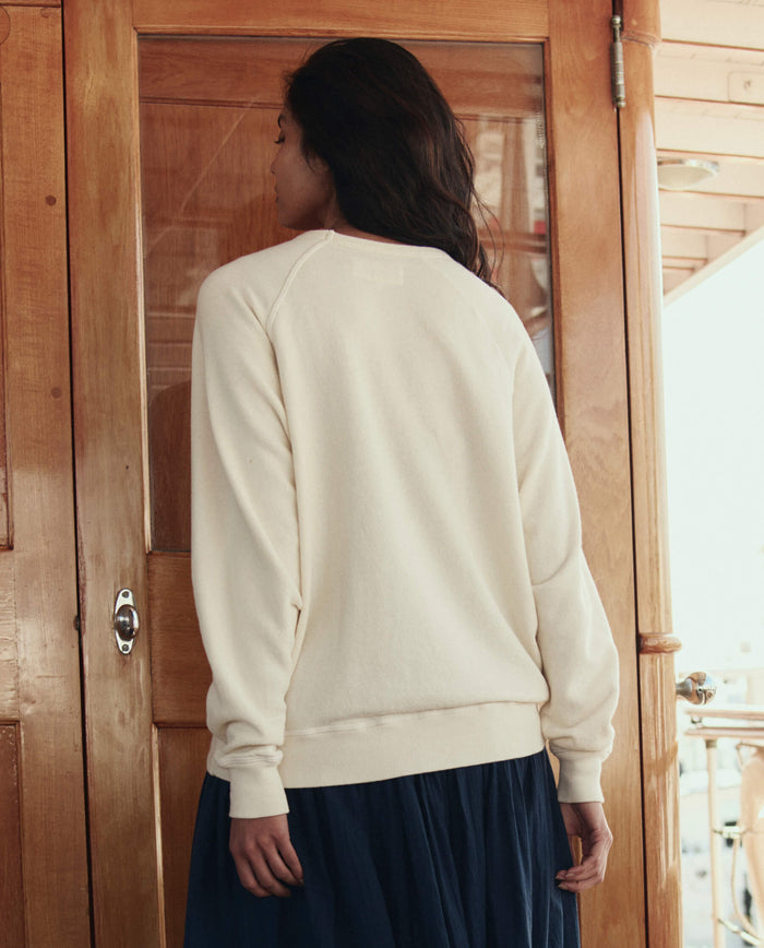 A person stands with their back to the camera, wearing THE GREAT. SU26 The College Sweatshirt W/ Sailing Boat Applique in Washed White and a dark skirt, in front of a wooden door.