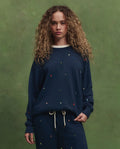 A woman with curly hair stands against a green background, wearing THE GREAT. PS26's The College Sweatshirt—Navy with Tossed Floral Embroidery—and matching sweatpants, both featuring delicate embroidered details.