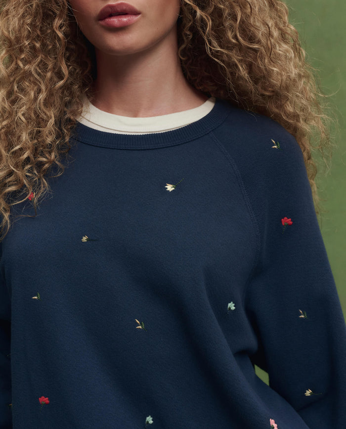 A person with curly hair wears THE GREAT. PS26 The College Sweatshirt in navy, featuring tossed floral embroidery, shown from shoulder to chest against a green background.