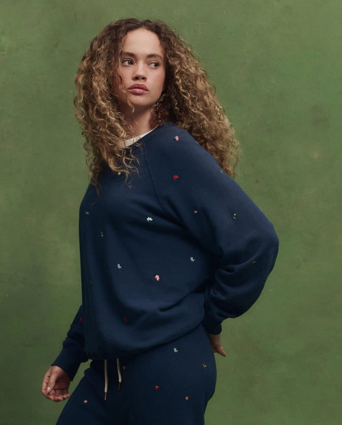 A person with curly hair wears THE GREAT. PS26 The College Sweatshirt in navy with tossed floral embroidery, paired with matching pants, standing before a green background and looking to the side.