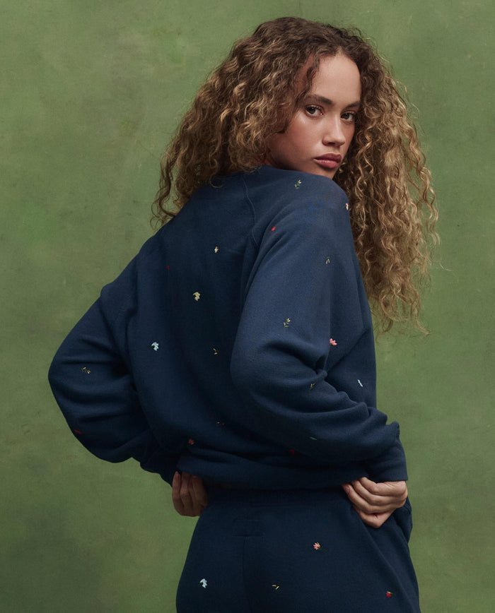 A person with curly hair wears THE GREAT. PS26 The College Sweatshirt, Navy with Tossed Floral Embroidery, and matching pants, standing against a green background and looking over their shoulder.