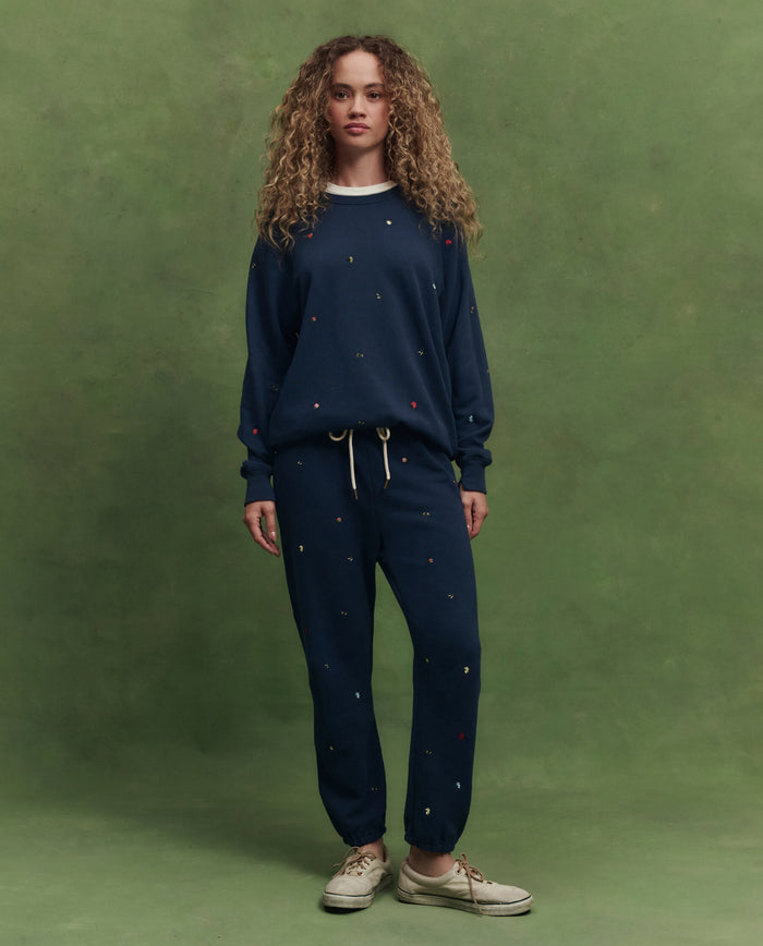 Against a green background, a person with curly hair wears THE GREAT. PS26’s College Sweatshirt in navy with tossed floral embroidery, paired with matching sweatpants and white sneakers.