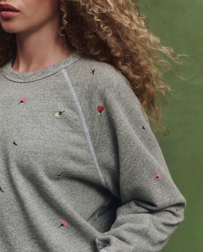 A person with curly hair wears THE GREAT. SU26 College Sweatshirt W/ Tossed Floral Emb. in Varsity Grey, featuring small, colorful floral embroidery, set against a green background.
