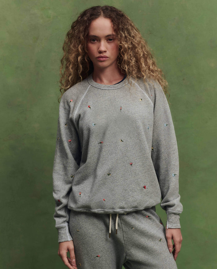A person with curly hair wears THE GREAT.’s The College Sweatshirt w/ Tossed Floral Embroidery in Varsity Grey, standing in front of a green background.