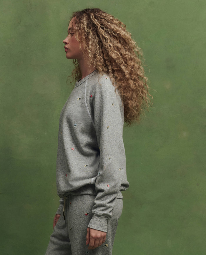 A person with curly hair in profile wears THE GREAT. SU26’s The College Sweatshirt W/ Tossed Floral Emb. in Varsity Grey, paired with matching pants, standing against a plain green background.