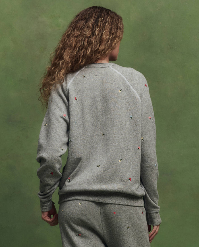 A person with curly hair, seen from the back, wears THE GREAT. SU26's The College Sweatshirt W/ Tossed Floral Emb. in Varsity Grey, featuring small colorful details, standing against a green background.