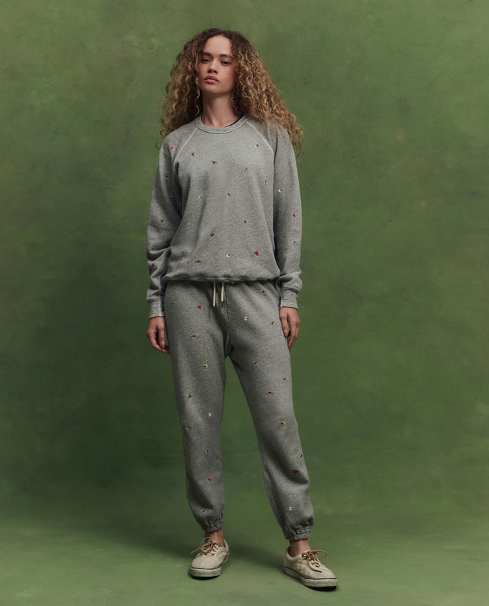Someone with curly hair wears THE GREAT. SU26 The College Sweatshirt W/ Tossed Floral Emb. in Varsity Grey, standing before a green backdrop, paired with matching sweatpants featuring small designs.