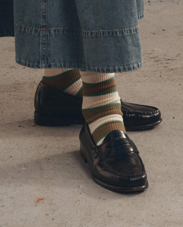 A person wearing THE GREAT. F26 D1 The Collegiate Sock. -- Green paired with black loafers and wide-leg blue jeans, standing on a concrete floor.