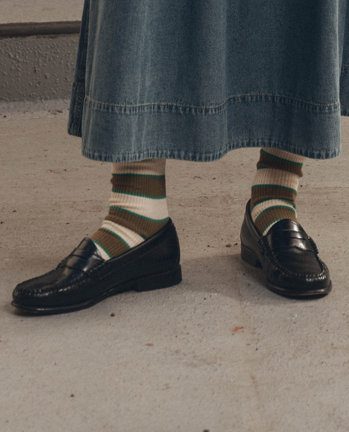 A person wearing a denim skirt, The Collegiate Sock in green by THE GREAT. F26 D1, and black leather loafers, standing on a concrete surface.