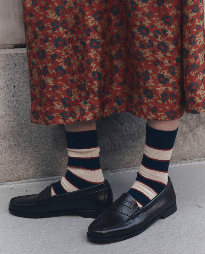 A person wearing black loafers, THE GREAT. F26 D1 The Collegiate Sock. -- Navy, and a red floral patterned skirt stands on a concrete surface.