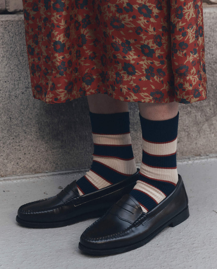 A person wearing a floral red skirt, THE GREAT. F26 D1 The Collegiate Sock. -- Navy in striped cream and dark blue, and black leather loafers standing on concrete.