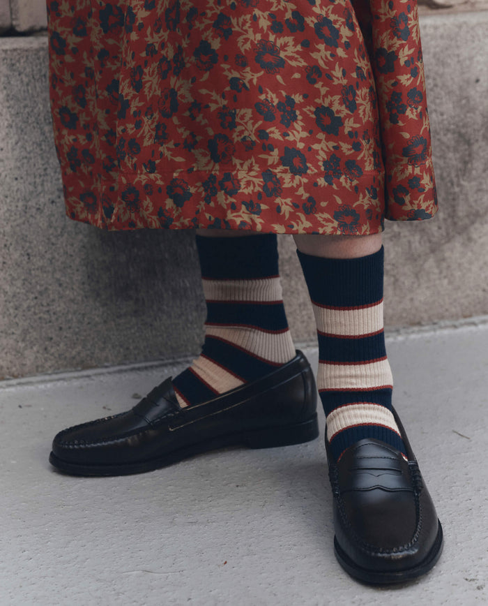 Person wearing THE GREAT. F26 D1 The Collegiate Sock. -- Navy, paired with black loafers and a long red floral skirt, standing on a light concrete surface.